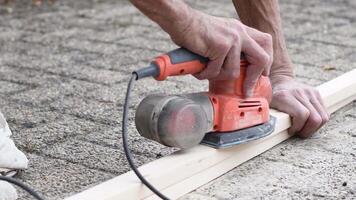 Builder male grinds a block of wood with a manual sandpaper electric grinder from bumps. Close Up view processes wooden beam by grinding, video