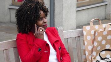 a young african american woman in a red coat sits on a bench after shopping and waits, angry at her husband who is late, she uses a smartphone to call him, video