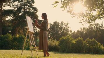 Créatif femme artiste des peintures une image avec une brosse tandis que permanent près le chevalet dans la nature. femme artiste, art pour ventes, inspiration dans la nature. video