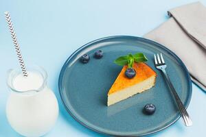 Slices of blueberry cake with mirror glaze decorated berries and leaves of mint with milk on a blue plate are on blue table, close up. photo