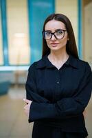 Pretty woman in glasses. Beautiful female in dark shirt stands cross hands and looks at camera. Closeup. photo