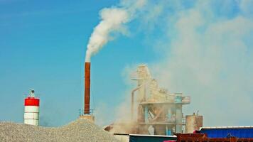 white steam rising up from pipe of small asphalt factory at day time with piles of gravel in a foreground video