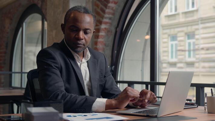 African American middle-aged entrepreneur typing laptop computer business  man office businessman male guy working with pc online job check data in  papers research analyse colourful diagrams documents