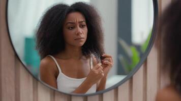 Worried beautiful sad upset African American woman girl female lady looking at mirror reflection holding fragile curly hair damaged dry curls unhealthy split ends hormone problem vitamins deficiency video