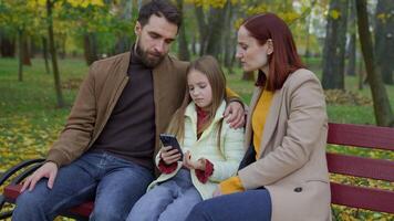 caucásico familia a ciudad banco juntos al aire libre otoño triste trastornado confuso preocupado con móvil teléfono malo Noticias teléfono inteligente fracaso pequeño hija niña niño niño con padres madre mujer padre hombre video