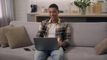 Happy smiling African American ethnic man working at home distant online work studying at sofa guy laptop using computer at couch male using pc playing internet game use app shopping chatting video