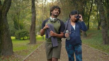 zwei Sport Menschen lächelnd reden Gehen zusammen im Park Stadt nach Laufen sich unterhalten Konversation Kommunikation freundlich indisch Frau Mädchen halten Wasser Flasche arabisch Mann Kerl mit Handtuch und Kopfhörer video