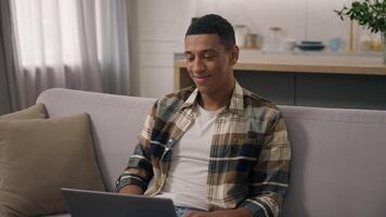 Happy smiling African American ethnic man freelancer working at home distant online work at sofa smile positive student guy studying remote with laptop using computer e-learning at couch male using pc video