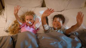 Funny playful laughing Caucasian children kids boy girl brother sister siblings happy family lying in bedroom on bed before bedtime waving hello greeting playing fooling around covered with blanket video