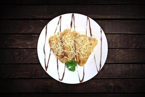 Three fried bananas topped with cheese and chocolate served on a white plate with a dark wood backdrop. photo