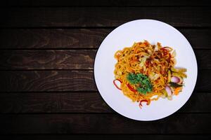 Spicy fried noodles with various additions on white plate on dark wooden table. top view. photo