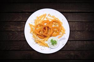 Onion rings and fries with tartar sauce served on a white plate on a dark table. photo