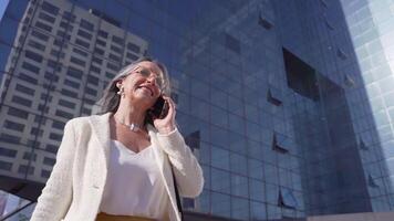 gelukkig aantrekkelijk zakenvrouw pratend met haar slim telefoon in voorkant van een modern gebouw. video