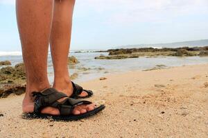 A pair of feet wearing black footwear is standing on the beach sand. photo