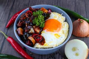 Rice in a bowl with sunny side up and fried chicken with black pepper sauce. Wooden background, chilies and onions. photo