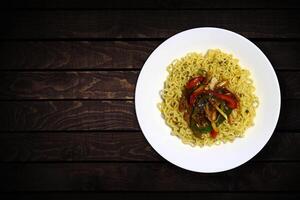 Top view of instant noodles in white bowl with added chopped vegetables and black peppers on dark wooden background. photo