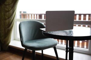 de cerca de un mesa con un ordenador portátil y un silla en frente de un grande ventana. remoto trabajo concepto foto