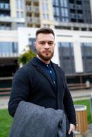 Cute man looks to the camera while wearing formal suit and holding gray coat in hands. Modern building on the background. photo