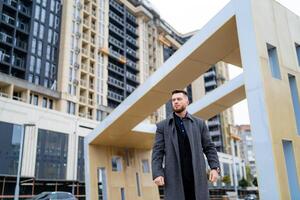 Trendy handsome man in gray coat and dark suit poses on modern architecture background. Beautiful photo zone.