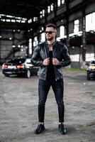 Stylish handsome man with sunglasses in fashionable clothes stands with the luxury black car on the background. Closeup. photo