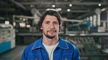 retrato de un joven trabajador en un espacioso taller a un fabricación planta. un simpático hombre en un azul uniforme mira a el cámara. video