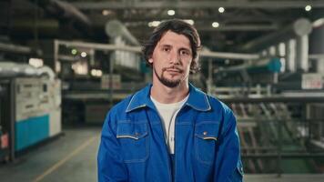 retrato de un joven trabajador en un espacioso taller a un fabricación planta. un simpático hombre en un azul uniforme mira a el cámara. video