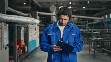 ein männlich Ingenieur im ein Blau Uniform Spaziergänge durch das Fabrik Werkstatt und prüft das Betriebs Parameter von ein automatisch Produktion Linie. Arbeit auf Rahmen oben automatisch Ausrüstung. video