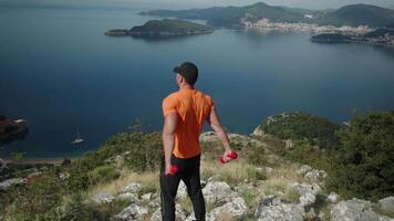 masculino atleta trenes espalda con pesas en naturaleza con hermosa mar puntos de vista video