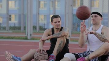 dos chicos son sentado en el baloncesto Corte con pelotas y comunicar. girar el pelota en su dedo. video