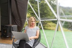 Woman working on laptop geo dome tents. Green, blue background. Cozy, camping, glamping, holiday, vacation lifestyle concept. Outdoors cabin, scenic background. photo