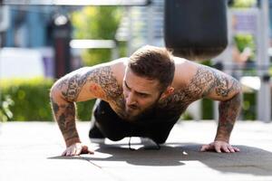 Muscular bodybuilder handsome man with naked torso trains triceps in a crossover cable exercise machine on the street photo