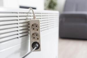 Electric heater plug inserted into extension cord. White radiator for home and office. House heating and comfort photo