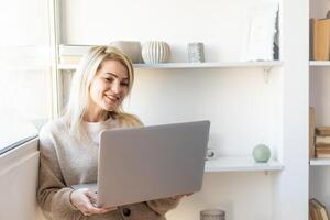 bella joven con ropa informal usando una laptop y sonriendo mientras trabaja en el interior foto