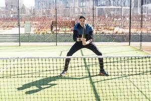 un padel jugador saltar a el pelota, bueno mirando para publicaciones y póster. hombre con negro raqueta jugando un partido en el abierto detrás el red Corte al aire libre. profesional deporte concepto con espacio para texto foto