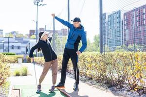 Sharing with golf experience. Cheerful young man teaching his daughter to play mini golf at the day time. Concept of friendly family photo