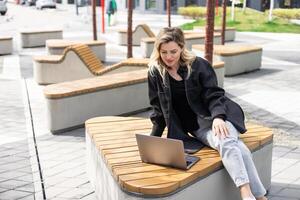 joven mujer sentado en en línea reunión en al aire libre cafetería, hablando a ordenador portátil cámara, explicando algo, Bebiendo café. foto