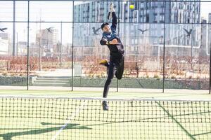 un padel jugador saltar a el pelota, bueno mirando para publicaciones y póster. hombre con negro raqueta jugando un partido en el abierto detrás el red Corte al aire libre. profesional deporte concepto con espacio para texto foto