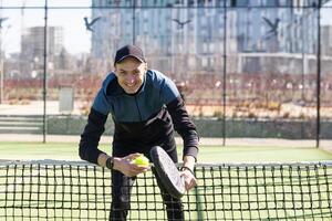 un padel jugador saltar a el pelota, bueno mirando para publicaciones y póster. hombre con negro raqueta jugando un partido en el abierto detrás el red Corte al aire libre. profesional deporte concepto con espacio para texto foto