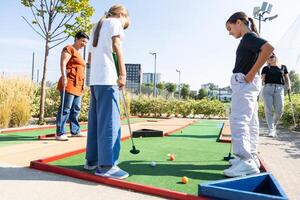 Cute school girl playing mini golf with family. Happy toddler child having fun with outdoor activity. Summer sport for children and adults, outdoors. Family vacations or resort. photo