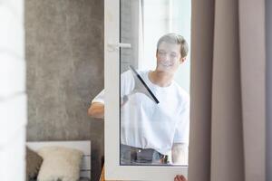 Side view of smiling man with bottle of detergent and rag cleaning window at home photo