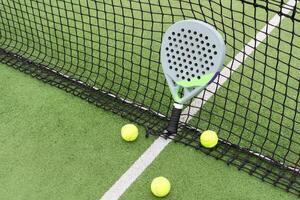 Yellow balls on grass turf near padel tennis racket behind net in green court outdoors with natural lighting. Paddle is a racquet game. Professional sport concept with copy space. photo