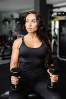 Young fit woman practicing dumbbell press while sitting on a bench. Slim female in sportswear exercises with weights in a gym photo