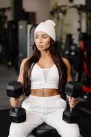 atlético joven aptitud mujer es vistiendo en blanco deporte traje en gimnasio atuendo haciendo hombro rutina de ejercicio con pesas foto