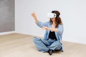 Young woman in VR glasses using modern technology creating room design, moving hands in air, sitting in new apartment during renovation. Virtual reality and repair concept. photo