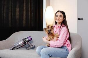 A cute young woman is sitting on a sofa with her dog, and a cordless vacuum cleaner with a sofa attachment is next to it photo
