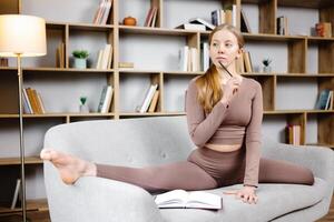 Young beautiful blonde woman in brown leggings and top sitting on a twine at home on the couch. The girl makes notes in a notebook while sitting on the sofa in an unusual position photo