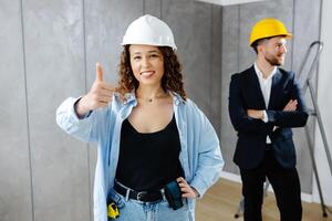 We've got the go ahead to renovate. Attractive young contractor and customer standing in room in helmet, looking at camera and showing gesture fingers up photo