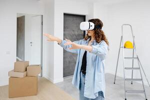 Young woman in VR glasses using modern technology creating room design, moving hands in air, standing in new apartment during renovation. Virtual reality and repair concept photo