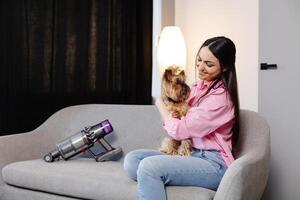 A cute young woman is sitting on a sofa with her dog, and a cordless vacuum cleaner with a sofa attachment is next to it photo