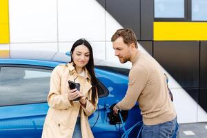 Young couple man and woman traveling by electric car having stop at charging station. Boyfriend plugging in cable to charge. Man talking with girlfriend, holding cup drinking hot coffee smiling photo